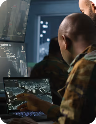 Two men in camouflage uniforms working on a laptop displaying a detailed map interface in a control room.